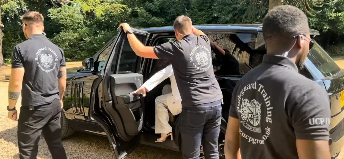 Close protection operatives in tactical gear undergoing Level 3 live training in an urban setting, with one operative providing cover while another escorts a VIP. UCP Group logo is visible on their uniforms.