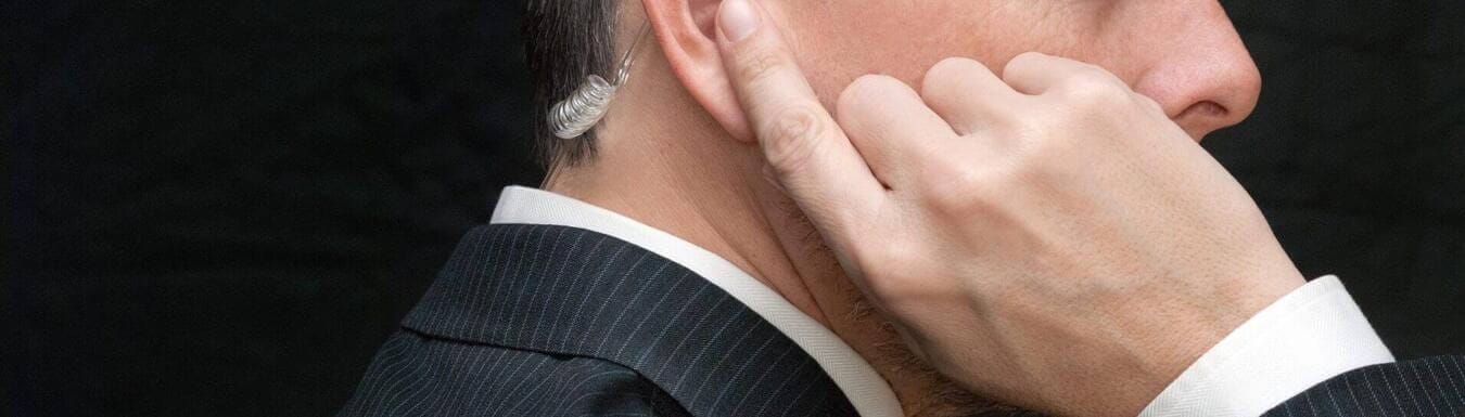 Close Protection Officer tapping Bluetooth earpiece, dressed in black tactical gear, staying alert on duty